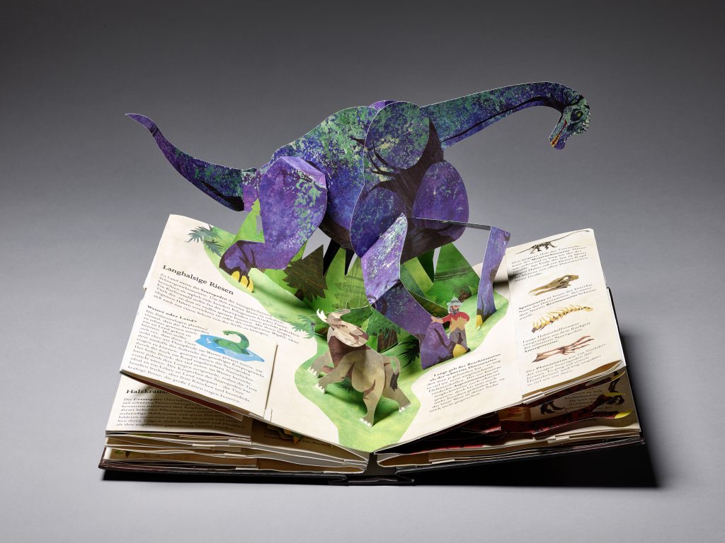 Ein aufgeklapptes Buch, aus dem sich durch das Aufklappen eine gefaltete Papierkonstruktion in Form eines langhalsigen Dinosauriers aufgestellt hat.