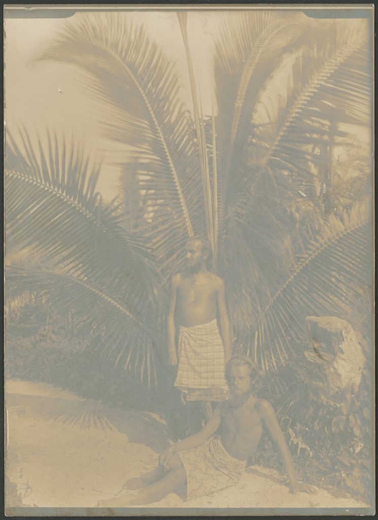Ein stehender und ein sitzender Mann, beide mit Lendenschurz bekleidet, vor einer Palme.