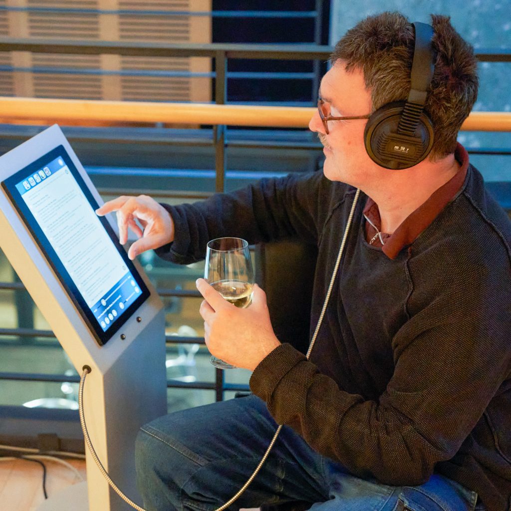 Ein Mann mit Kopfhörern und einem Weinglas in der Hand sitzt auf einem Sessel. Er schaut auf ein vor ihr stehendes Display und wählt etwas mit einem Finger aus. 