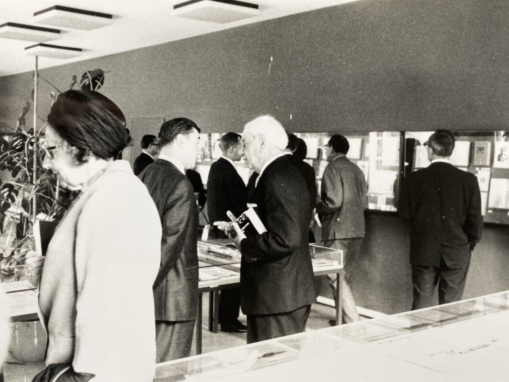 Schwarz-weiß Foto. Menschen schauen sich die Ausstellung an. Ein Mann hat den Ausstellungskatalog unter dem Arm.