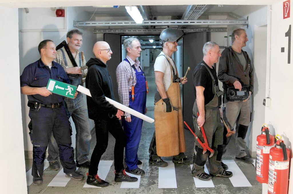 Sieben Haustechniker stehen mit ihren Werkzeugen im Kellerflur auf einem Zebrastreifen