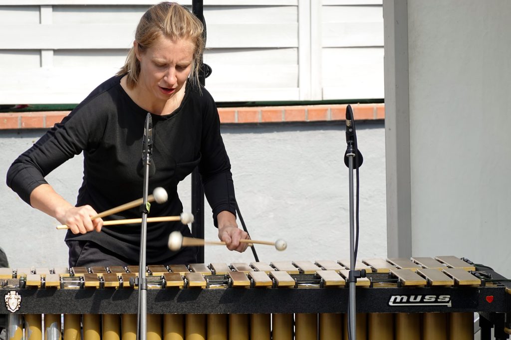 Els Vandeweyer, blonde Frau, schwarze Hose, schwarzes Oberteil, spielt am Vibraphone. Sie hält in jeder Hand je zwei Klöppel. Im Hintergrund ist eine Hausmauer zu sehen.