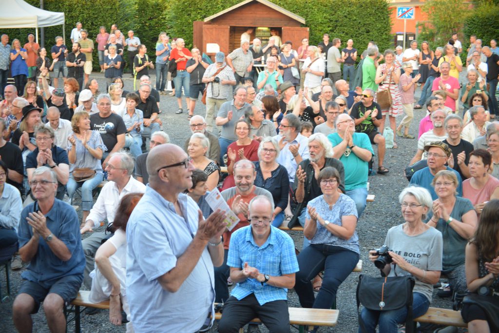 Ulli Blobel (Mann, Glatze, schwarze Brille, hellblaues halbarm Hemd)dahinter und um ihn herum Publikum auf Bierbänken und stehend.