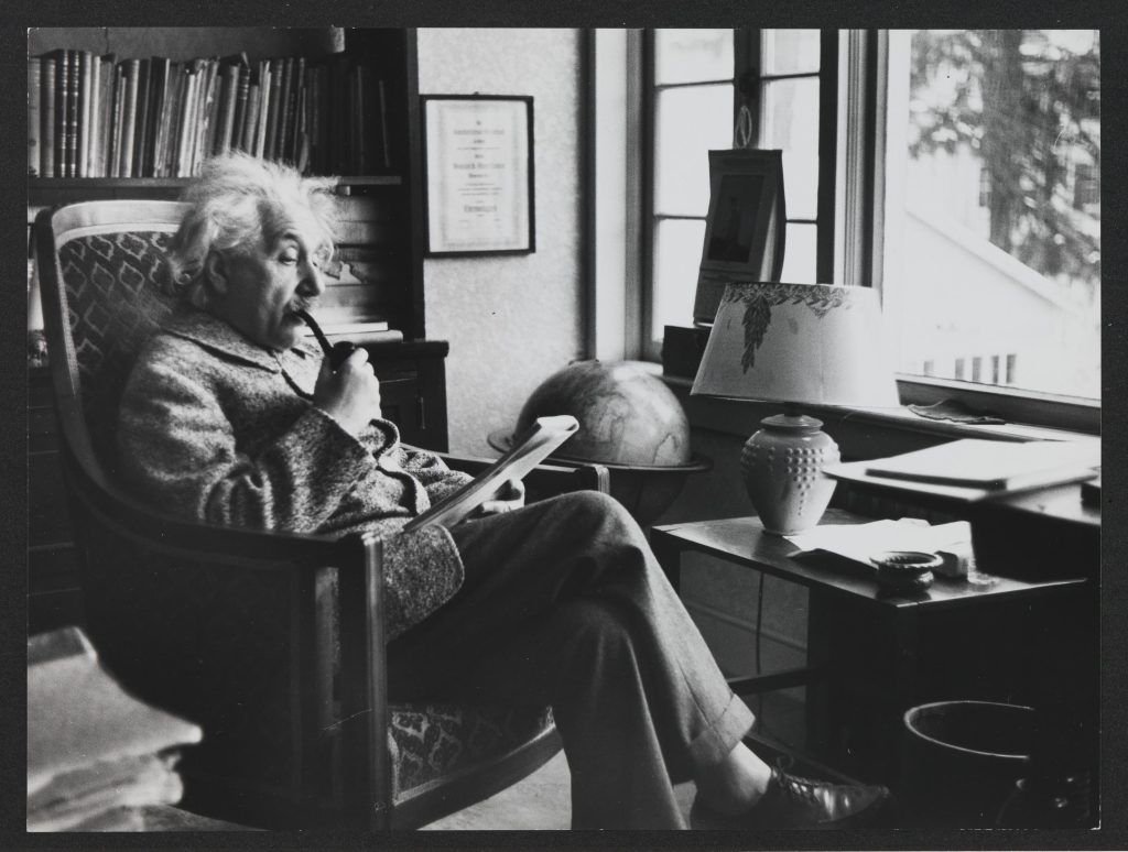 Albert Einstein 17.01.1939 in einem Zimmer seines Hauses in Princeton. Im Sessel sitzend, lesend vor Bücheregal und Fenster, die obligatorische Pfeife im Mund. Ein Bein über das andere geschlagen, ist am unteren Bildrand gerade noch der rechte Fuß, mit Lederslipper, ohne Socken sichtbar.