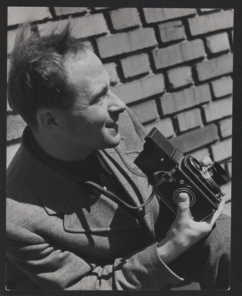 Eric Schaal im Profil vor einer Klinker-Mauer kniend/hockend. Er hält eine Kamera der 30er/40er Jahre in der Hand.