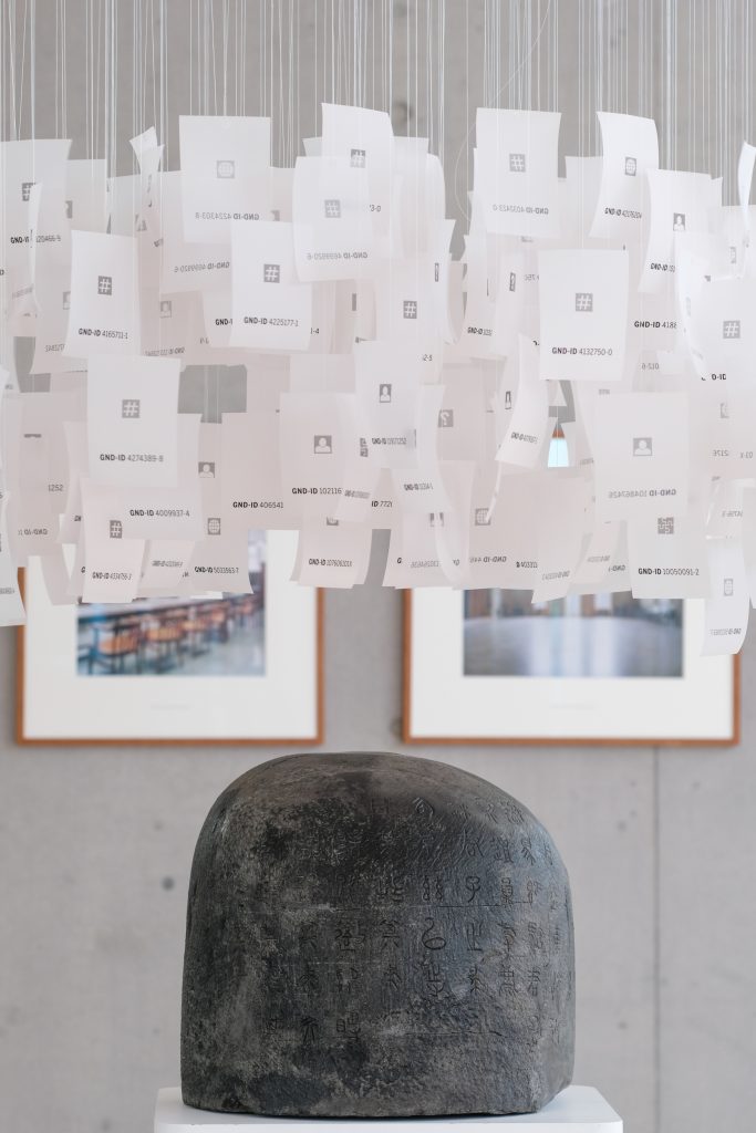 Foto eines runden Steinobjektes, darüber hängen Papierblätter