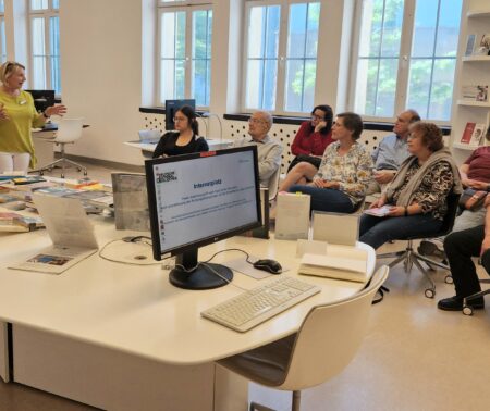 Eine Frau und ein Mann stehen vor einer sitzenden Gruppe Menschen in einem hellen, weiß gehaltenen Raum. Die Frau spricht zu der Gruppe.