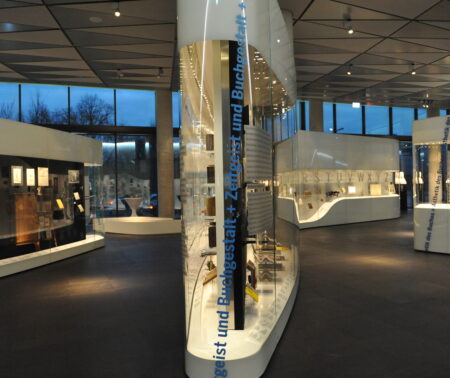 Mehrere beleuchtete Vitrinen. Auf der vordesten Vitrine steht der Schriftzug "Zeitgeist und Buchgestalt". Im Hintergrund sieht man die Dämmerung. Deutsches Buch- und Schritmuseum in d