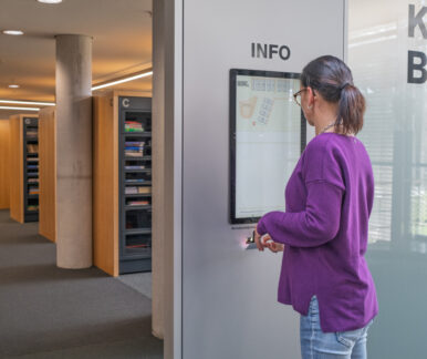 Im Zentrum des Bildes sieht man eine Informationsstele der Medienboxen. Sie ist mit Info beschriftet. Eine Frau scannt ihre Nutzer*innenkarte