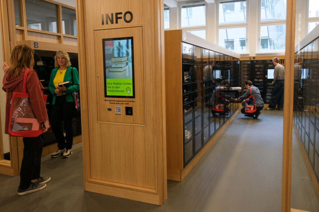 Im Zentrum des Bildes sieht man eine Informationsstele der Medienboxen. Sie ist mit Info beschriftet und zeigt ein Bild mit der Unterschrift "Ihre Medien finden Sie in den Medienboxen" Ringsherum nutzen Menschen die Medienboxen
