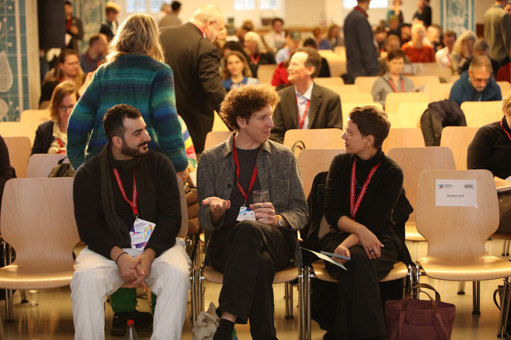 In der ersten Reihe sitzen drei Menschen, die sich unterhalten