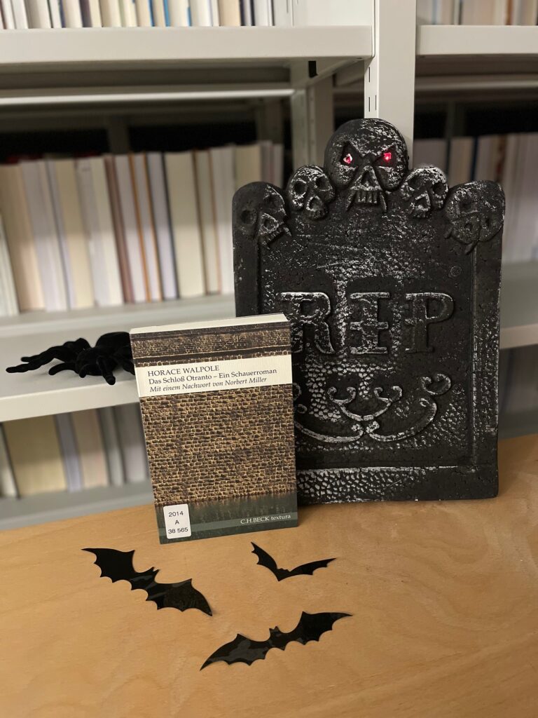 Buch Das Schloss von Otranto im Buchregal mit Deko-Grabstein, Plüschspinne und Plastikfledermäusen