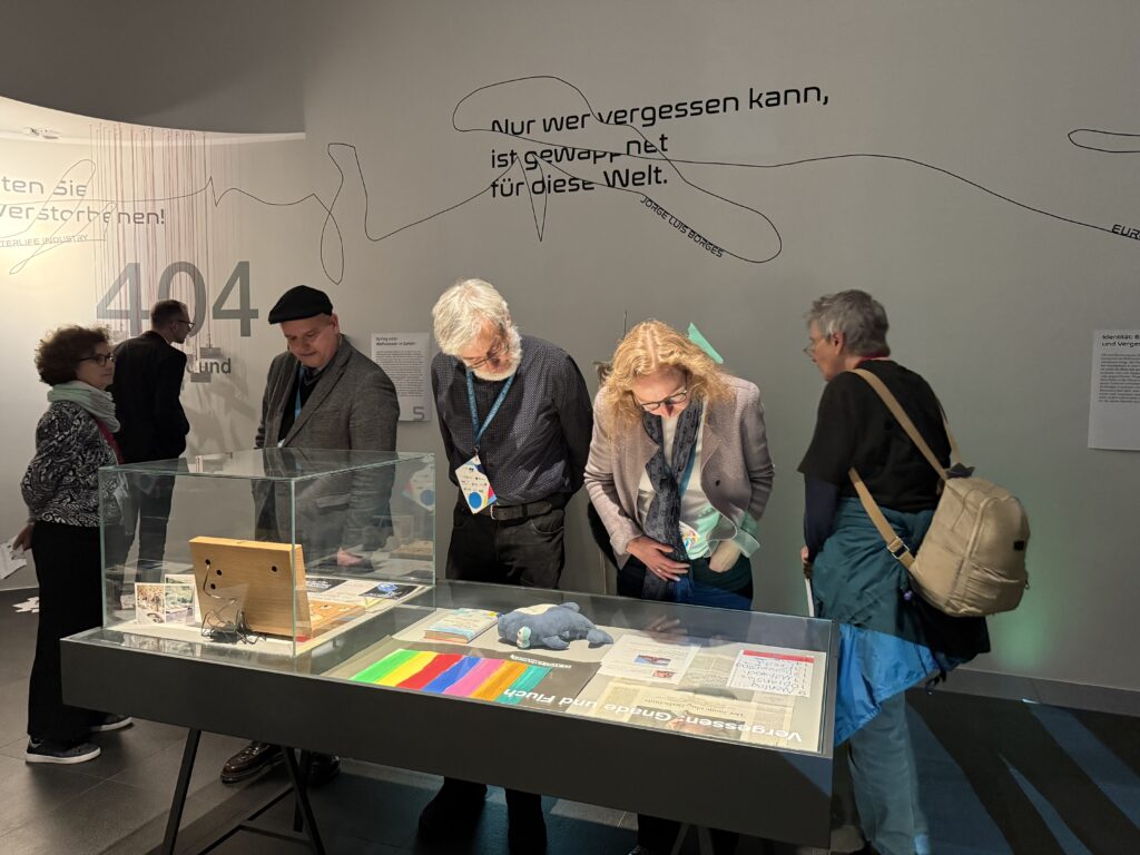 Mehrere Menschen schauen sich eine Vitrine in einer Ausstellung an. Als Objekte sind mehrere Papier sowie ein Kuscheltier-Hai zu sehen.