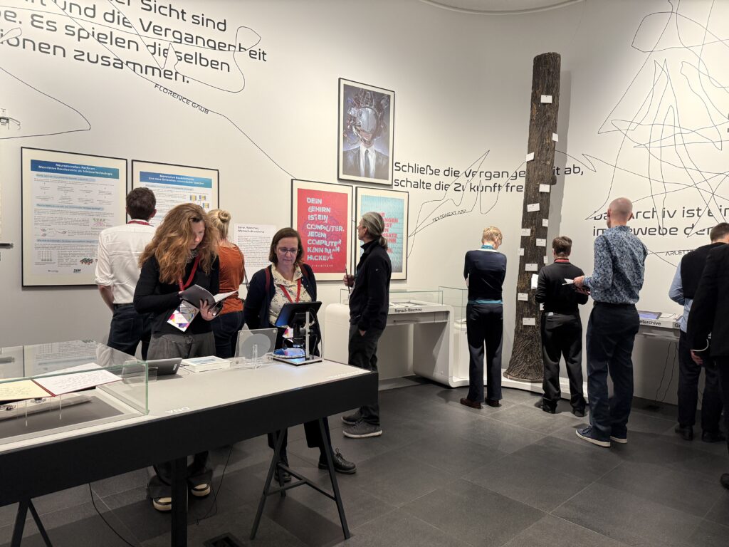 Viele Menschen schauen sich eine moderne Ausstellung an. Eine Frau hält ein Buch, eine andere schaut sich etwas auf einem Bildschirm an.