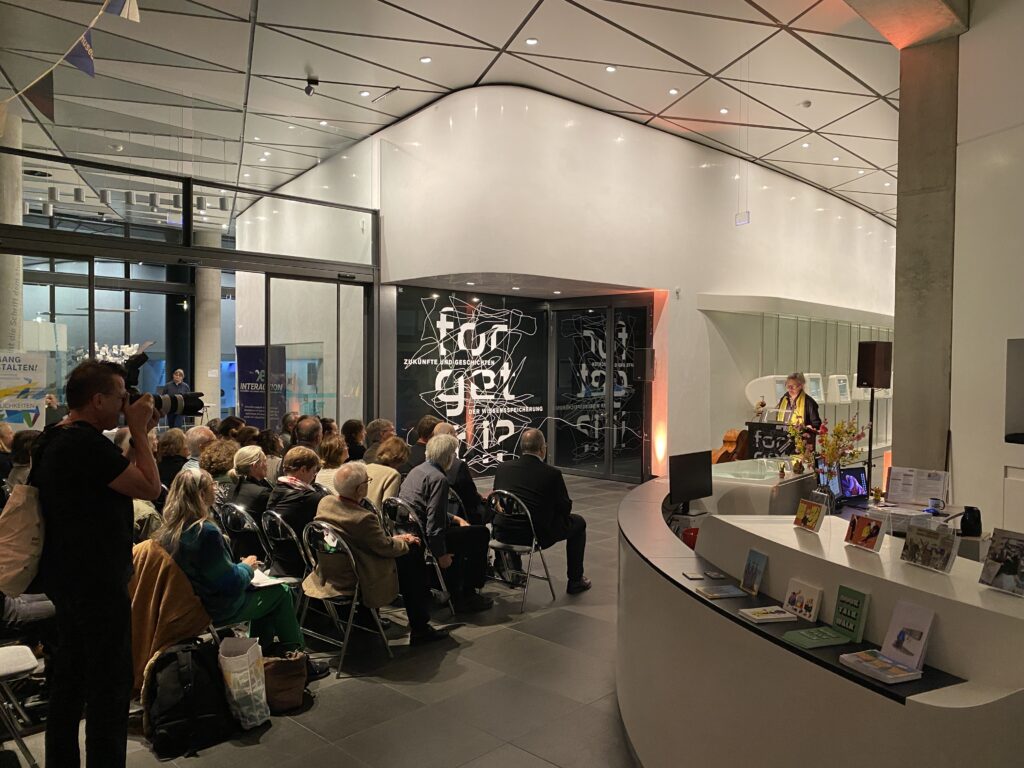 Eine Frau hält eine Rede in einem modernem Gebäude. Die Zuhörer sitzen in Reihen davor.