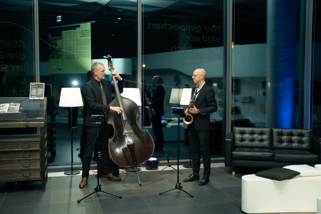 Zwei Musiker stehen vor einer Glasfront. Einer spielt Kontrabaß, der andere Saxophon