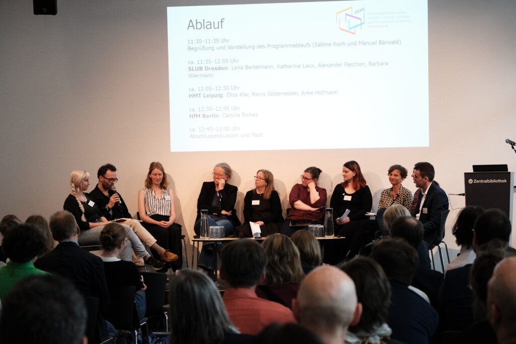 Menschen sitzen auf einer Bühne im Halbkreis. Eine Person redet. Die Menschen im Publikum vor der Bühne und auf der Bühne hören interessiert zu.
