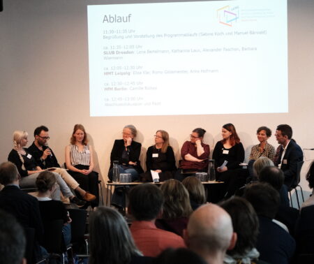 Menschen sitzen auf einer Bühne im Halbkreis. Eine Person redet. Die Menschen im Publikum vor der Bühne und auf der Bühne hören interessiert zu.