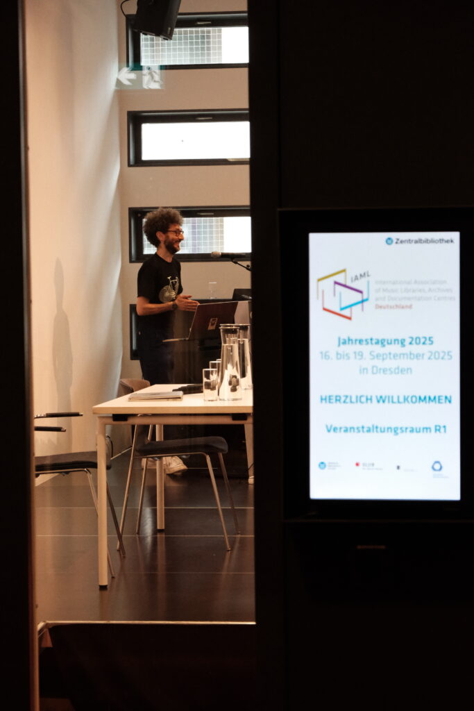 Eine fröhliche Person steht vor einem Pult und hält einen Vortrag. Sie ist durch die Scheibe einer Tür zu sehen.