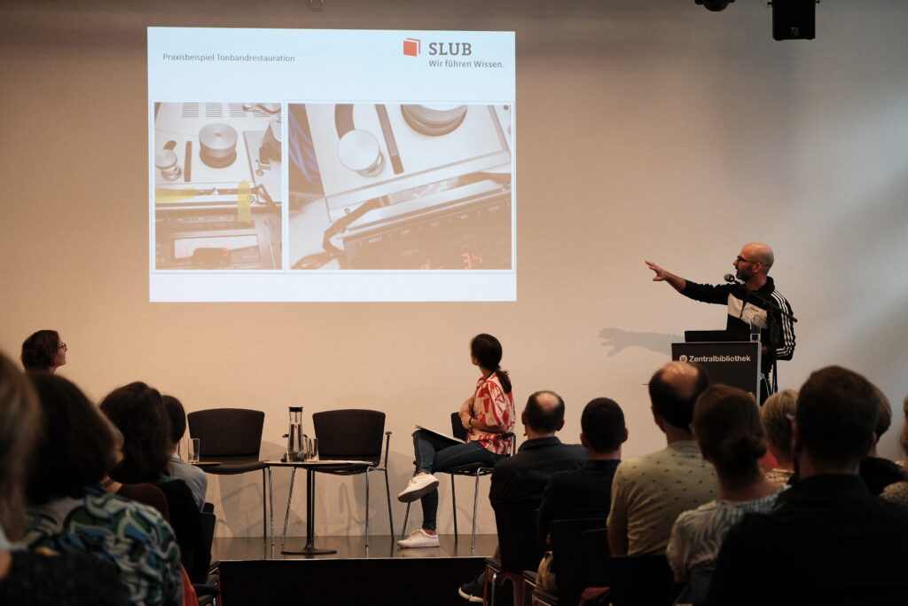 Viele Menschen sitzen im Publikum und hören jemandem zu, der einen Vortrag hält.