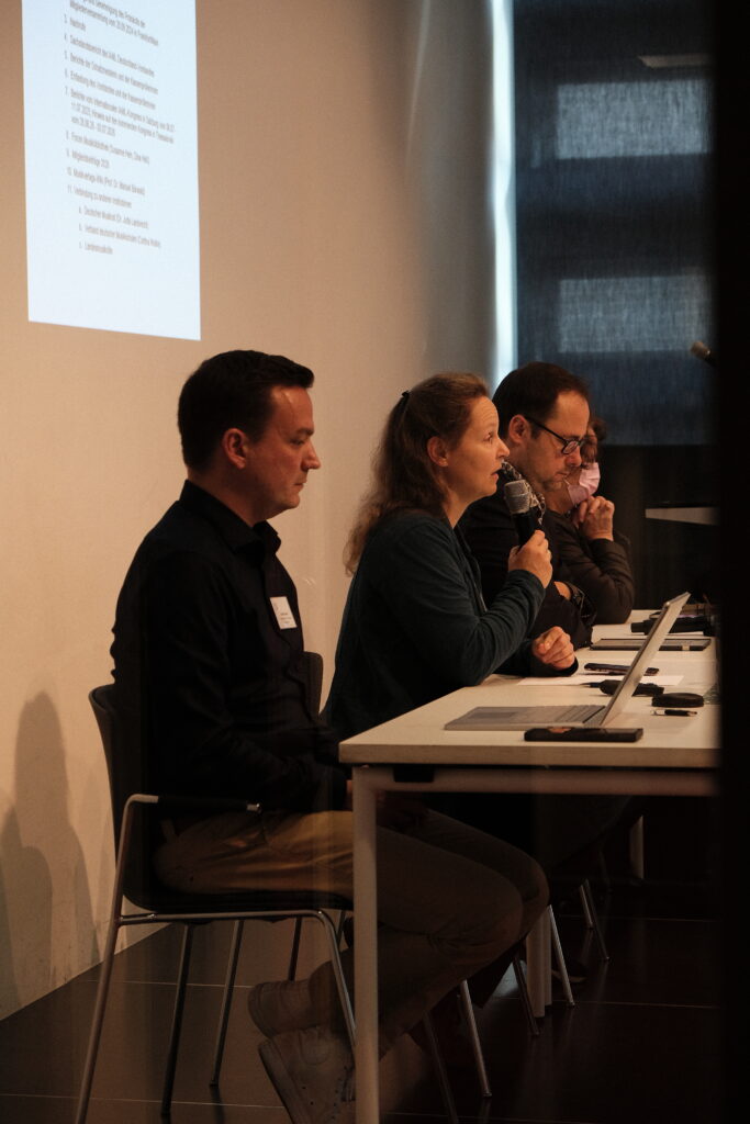Vier Personen sitzen an einem Tisch. Eine Person redet in ein Mikrofon während die anderen Zuhören.