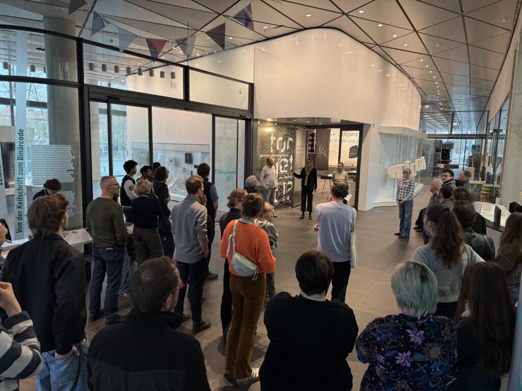 Viele Menschen stehen vor einer Ausstellung und hören einer Person zu, die eine Rede hält.