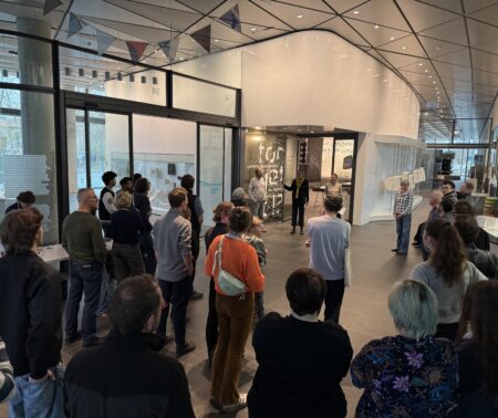 Viele Menschen stehen vor einer Ausstellung und hören einer Person zu, die eine Rede hält.