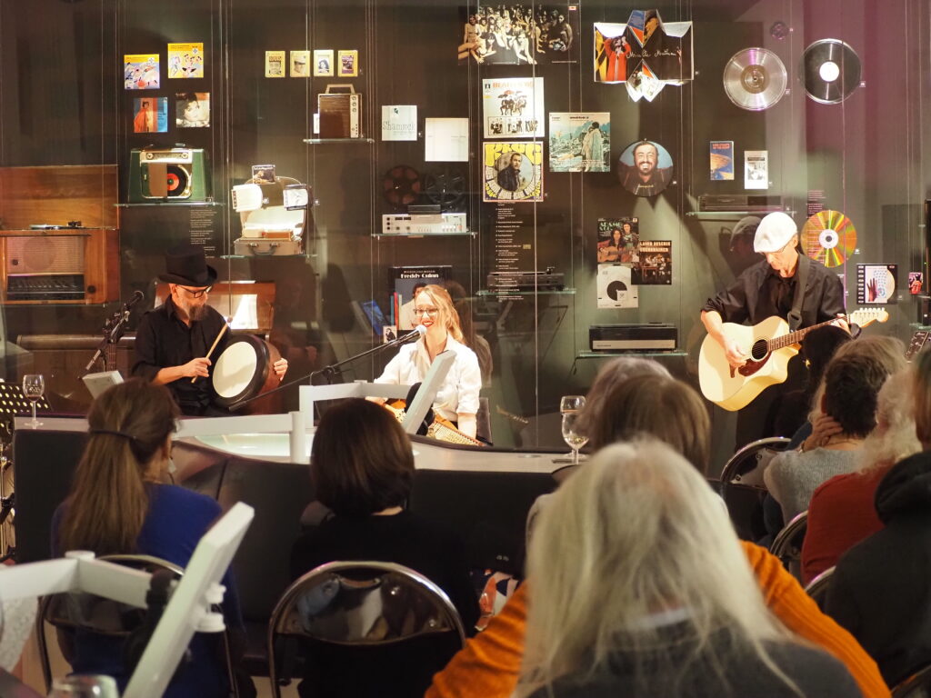 Die Band besteht aus drei Personen. Im Vordergrund sitzen Zuhörer.