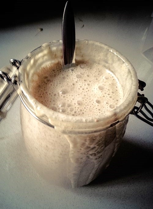 Glas mit schäumendem Sauerteig. Darin steckt ein Löffel.