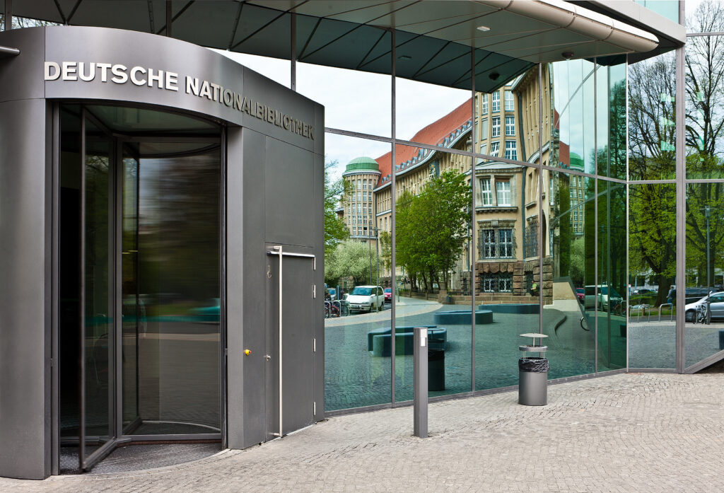 Man sieht eine dunkelgraue Drehtür in modernem Design. Sie ist mit dem Schirftzug "Deutsche Nationalbibliothek" überschrieben. Rechst neben der Tür befindet sich eine große Fensterfront, die aus mehreren schmäleren hochkantigen Fenstern zusammengesetzt ist. Dort sieht man die deutlich die Spiegelung eines historischen Gebäudes, aus beigen Sandstein. Es hat hervorstehende Erker über die vollständige Höhe des Gebäudes, in denen sich mehrere Sprossenfenster befinden. Zwischen den beiden Gebäuden verläuft eine Straße aus Kopfsteinpflaster.