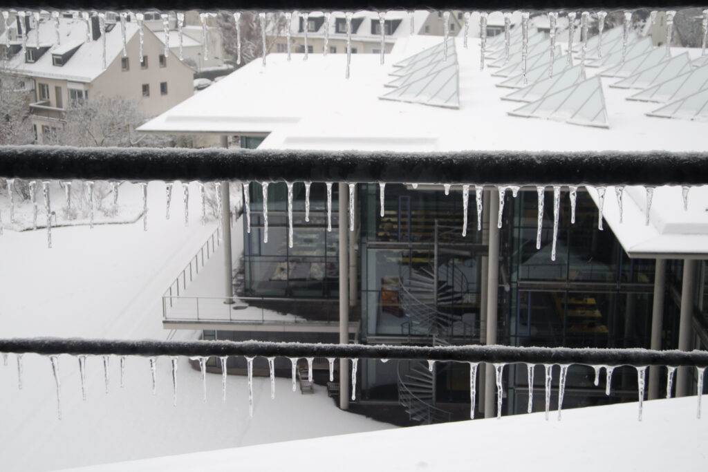 Man sieht eine städtische Umgebung im Schnee. Im Vordergrund sieht man die Querstreben eines Balkongeländers, an denen viele kleine Eiszapfen hängen. Man schaut durch sie hindurch auf ein Gebäude im Hintergrund. DIeses Gebäude hat große Glasfenster durch die man die Bücherregale im inneren sehen kann. Auf dem Dach des Gebäudes und auf der freien Fläche davor liegt Schnee.