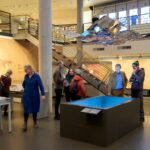 Besucherinnen und Besucher stehen in einer Ausstellungshalle um einen blauen, quadratischen Medientisch; darüber hängt eine spiegelnde Metallskulptur, im Hintergrund eine Treppe.