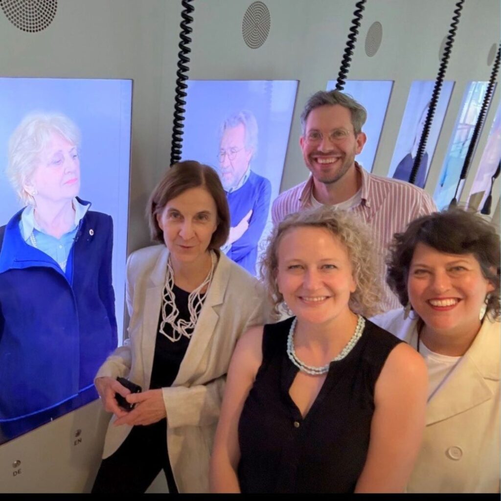 Vier Personen machen ein Gruppenfoto in einem Ausstellungsraum, hinter ihnen leuchten Bildschirme mit Porträts; Einhandkopfhörer hängen von den Bildschirmen herab.