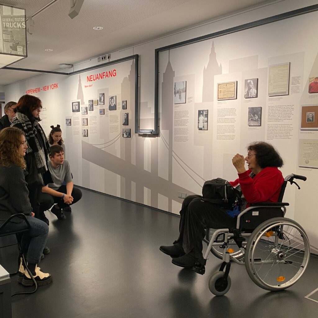 Eine Frau im Rollstuhl spricht zu einer kleinen Gruppe in einer Ausstellung; an der Wand hängen Texte und Fotos unter der Überschrift „Neuanfang“.