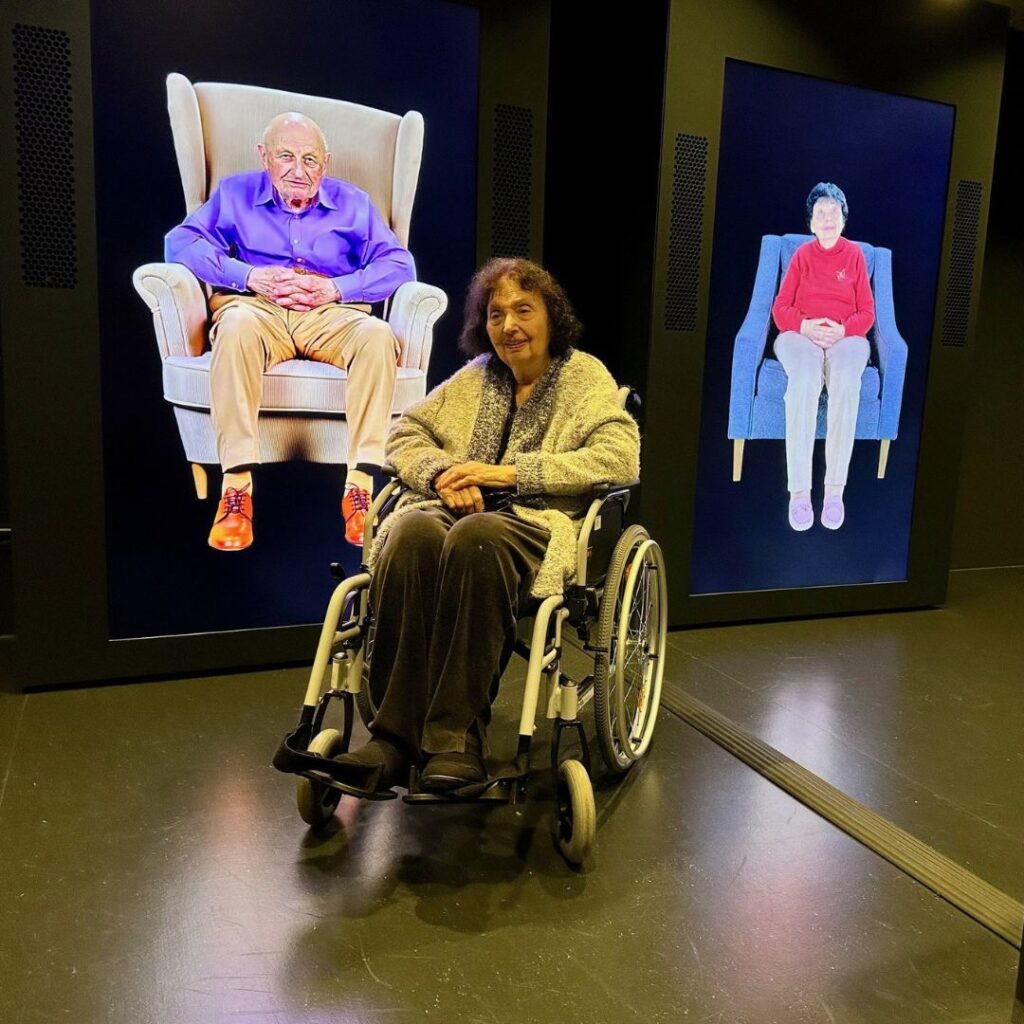 Eine ältere Frau im Rollstuhl sitzt vor zwei großen Videoporträts von Menschen in Sesseln in einem dunklen Ausstellungsraum.