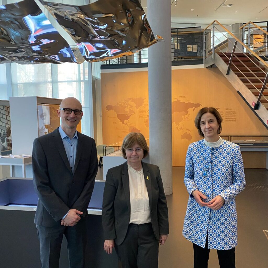 Drei Personen posieren in einem hellen Museumsfoyer unter einer großen, spiegelnden Metalllampe; im Hintergrund Treppe und Ausstellungsbereich.