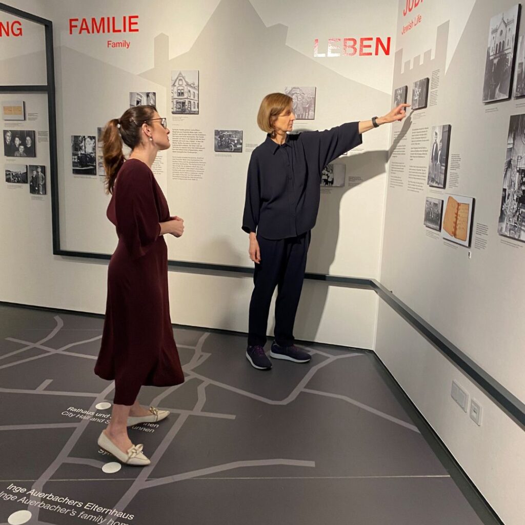 Zwei Frauen stehen in einer Museumsausstellung; eine zeigt auf eine Wand mit historischen Fotos und Texttafeln, der Boden ist wie eine Karte gestaltet.
