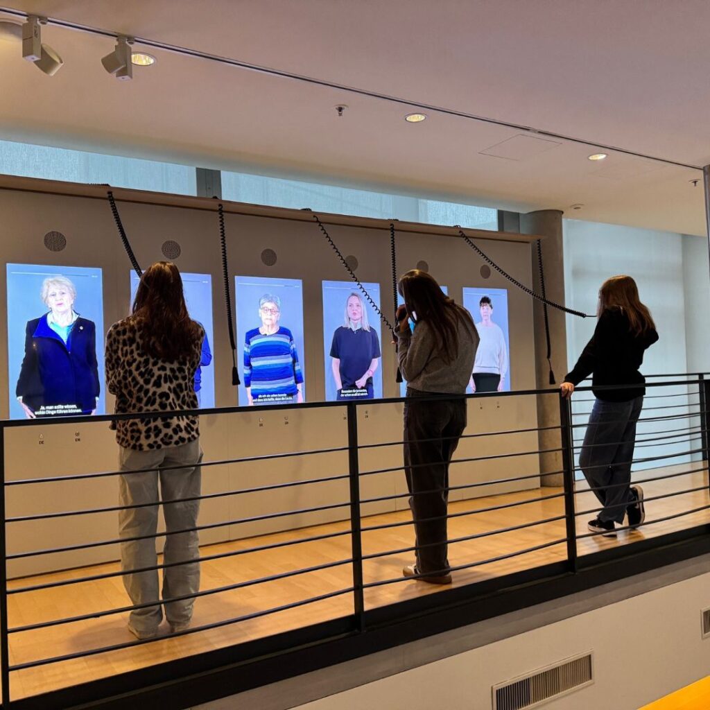 Drei Besucherinnen stehen an einem Geländer und hören über kabelgebundene Hörer Video-Interviews auf großen Bildschirmen.