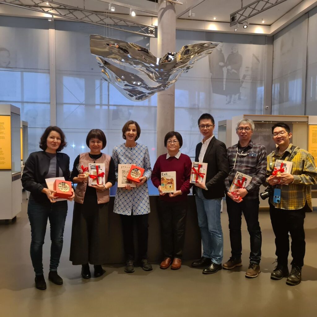 Sieben Personen stehen nebeneinander in einer Ausstellungshalle und halten Geschenkpakete und Bücher mit roten Schleifen; über ihnen hängt eine Metallskulptur.