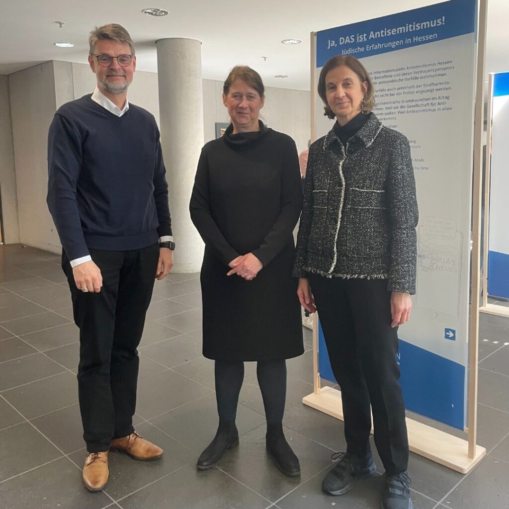 Drei Personen posieren in einem Foyer neben einer Infotafel mit der Überschrift „Ja, DAS ist Antisemitismus! Jüdische Erfahrungen in Hessen“.