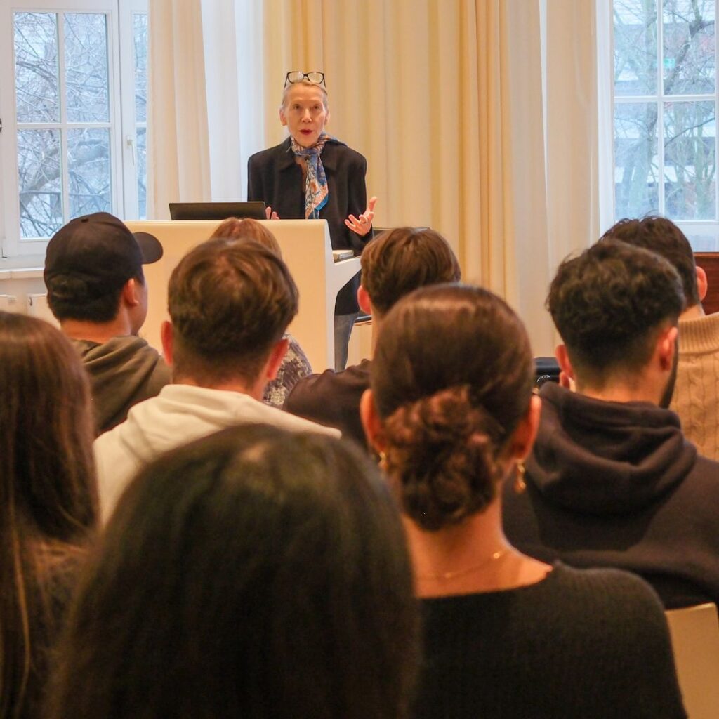 Vortragssituation: Eine Referentin spricht hinter einem Pult, das Publikum sitzt im Vordergrund und hört zu.