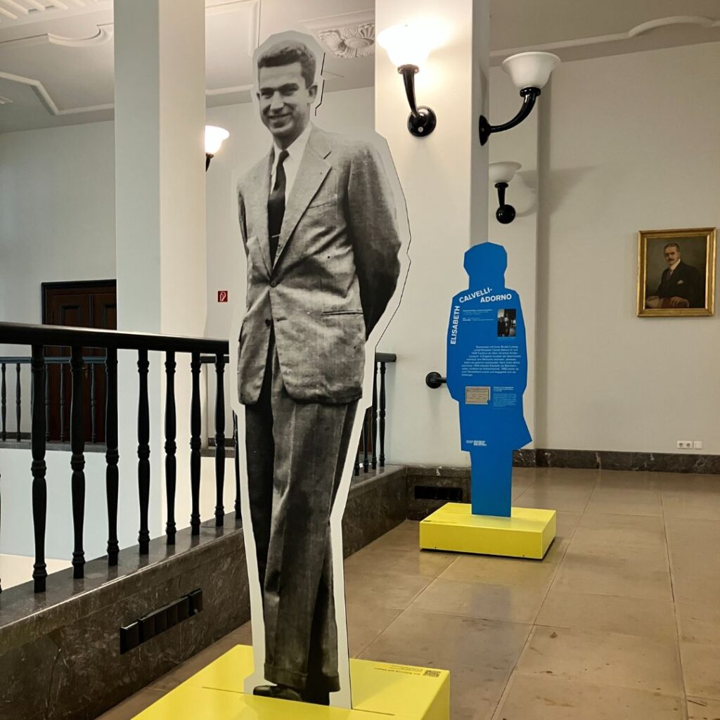 Flur mit lebensgroßem Schwarzweiß-Aufsteller eines Mannes im Anzug; daneben ein blaues Infodisplay in Form eines Menschen und ein gerahmtes Porträt an der Wand.