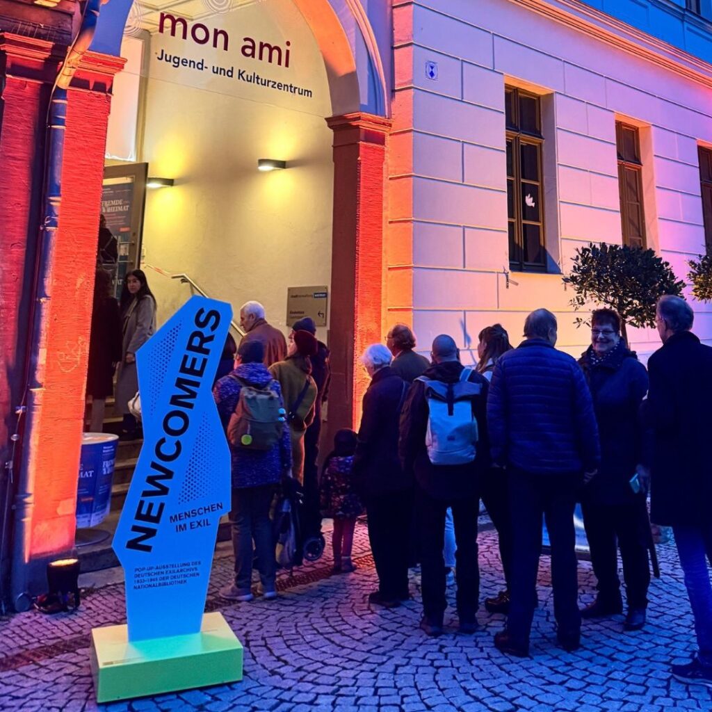 Abendliche Warteschlange vor dem Eingang des Kulturzentrums „mon ami“; im Vordergrund ein blaues Schild „NEWCOMERS Menschen im Exil“.