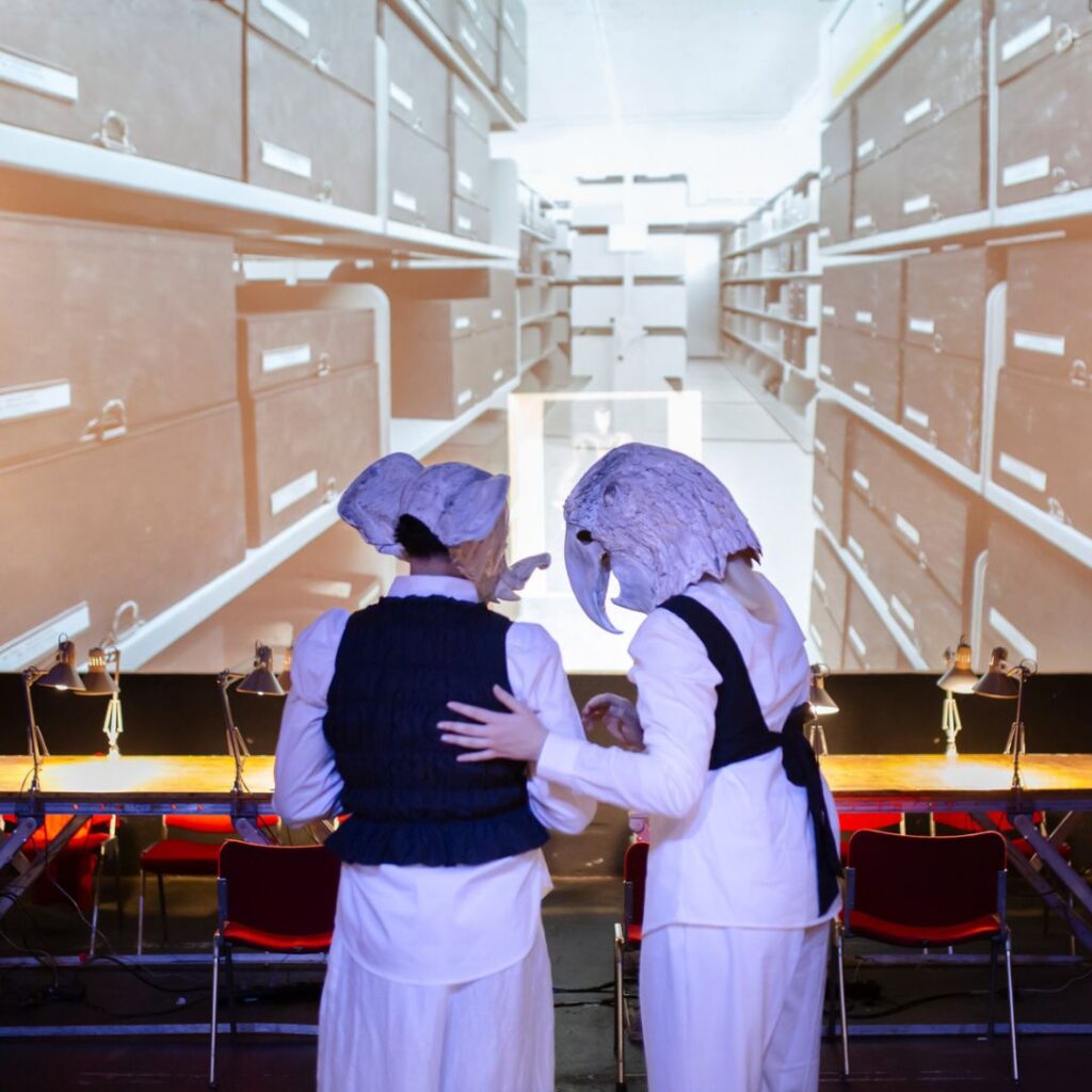 Zwei kostümierte Personen mit großen Masken stehen vor einer Projektion von Archivregalen, eine legt der anderen den Arm um die Schulter.