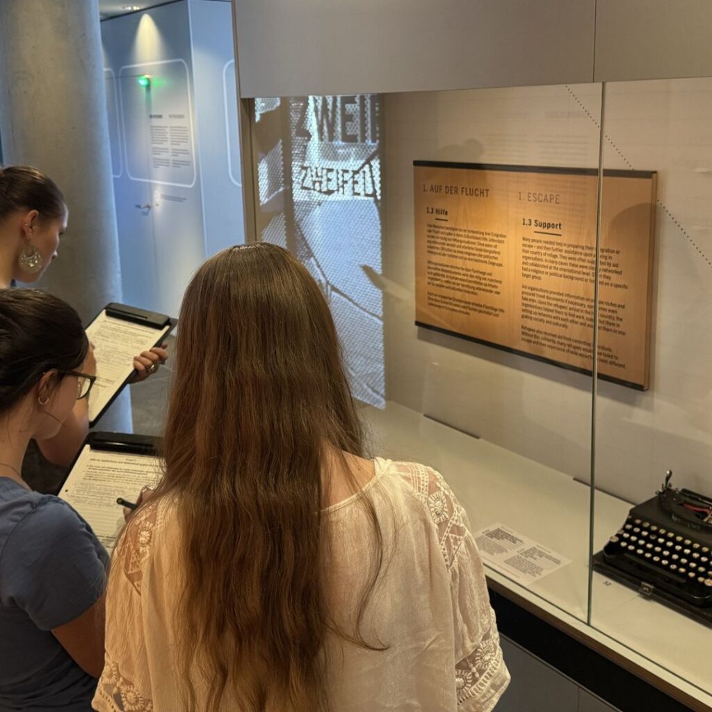 Drei Besucherinnen lesen auf Klemmbrettern vor einer Ausstellungsvitrine mit Schreibmaschine; dahinter eine Projektion und ein Textpanel „Auf der Flucht / Escape“.