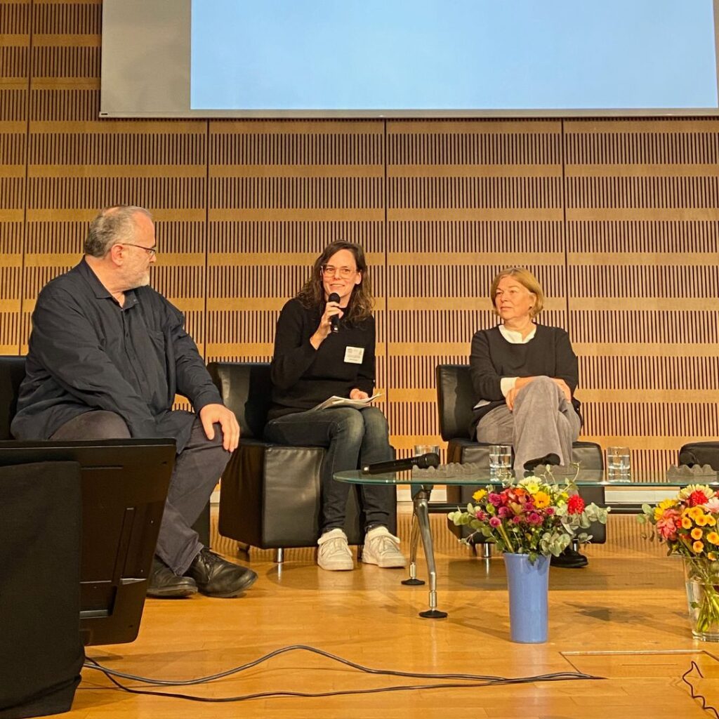 Podiumsgespräch auf einer Bühne: drei Personen sitzen auf Sesseln, eine spricht ins Mikrofon, davor Blumen und Wassergläser.