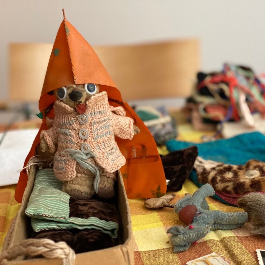 Nahaufnahme einer kleinen Stoffpuppe mit großer Kapuze und Strickmantel, sitzend in einer kleinen Schachtel wie in einem Bett; daneben liegen Miniatur-Stofftiere und Stoffteile.