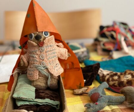 Close-up of a small teddy bear wearing a knitted coat and an orange raincoat, sitting in a small box like a bed; miniature stuffed animals and textile accessories lie beside it.