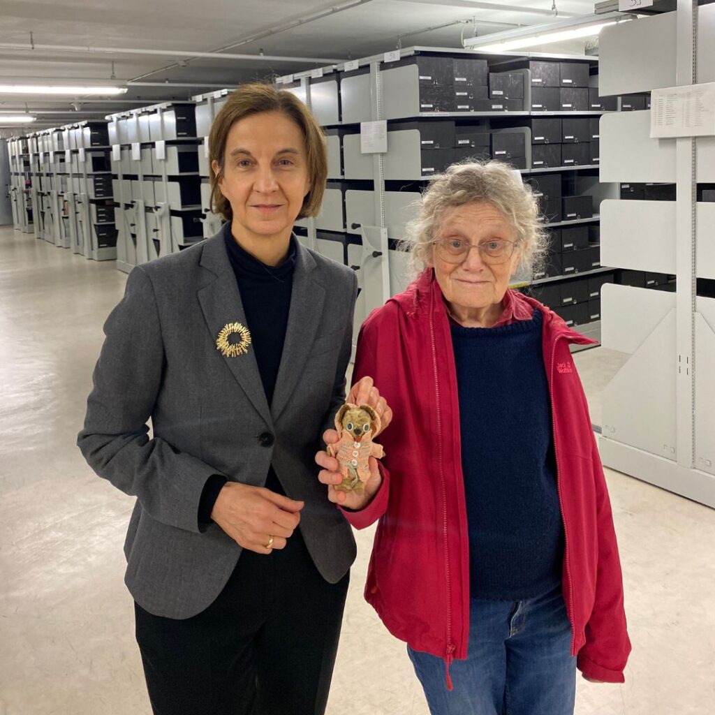 Zwei Frauen stehen in einem Archivmagazin zwischen langen Regalreihen mit Archivboxen; eine hält eine kleine Stoffpuppe in Strickkleidung in die Kamera.