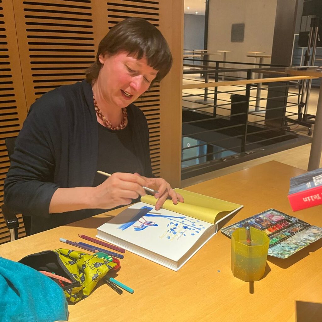 Eine Person illustriert mit Pinsel und Wasserfarben in einem offenen Buch; daneben liegen Stifte, Farbpalette und Wasserbecher auf dem Tisch.